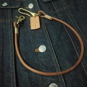 Puede incluir: Una cadena de cuero marr&oacute;n para cartera con herrajes de lat&oacute;n y una peque&ntilde;a etiqueta rectangular de cuero. La cadena est&aacute; en bucle y sujeta a una chaqueta vaquera, mostrando el dise&ntilde;o y la funcionalidad del accesorio.