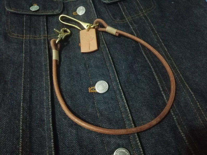 Puede incluir: Una cadena de cuero marr&oacute;n para cartera con herrajes de lat&oacute;n y una peque&ntilde;a etiqueta rectangular de cuero. La cadena est&aacute; en bucle y sujeta a una chaqueta vaquera, mostrando el dise&ntilde;o y la funcionalidad del accesorio.