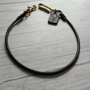 May include: A black leather cord lanyard featuring a brass swivel lobster clasp on one end and a brass hook on the other. A small rectangular black leather tag embossed with a tree silhouette is attached to the brass hook, completing the accessory.