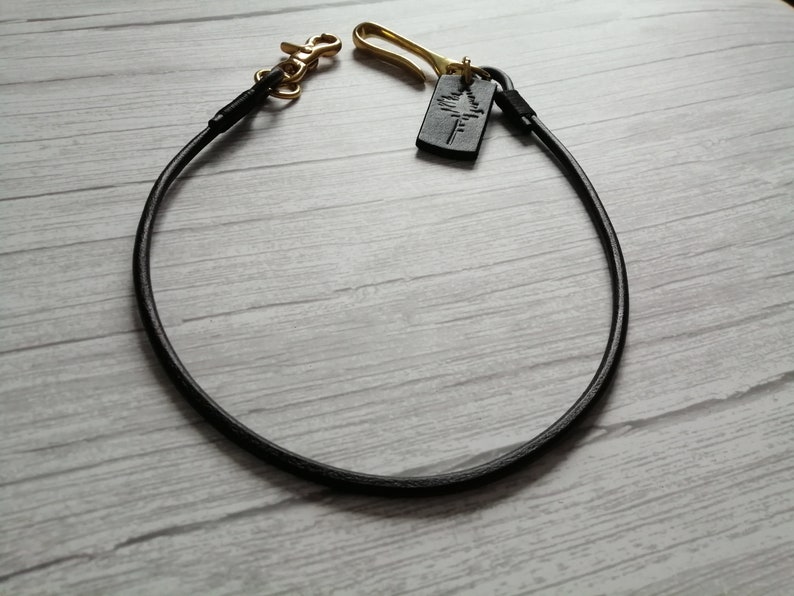 May include: A black leather cord lanyard featuring a brass swivel lobster clasp on one end and a brass hook on the other. A small rectangular black leather tag embossed with a tree silhouette is attached to the brass hook, completing the accessory.