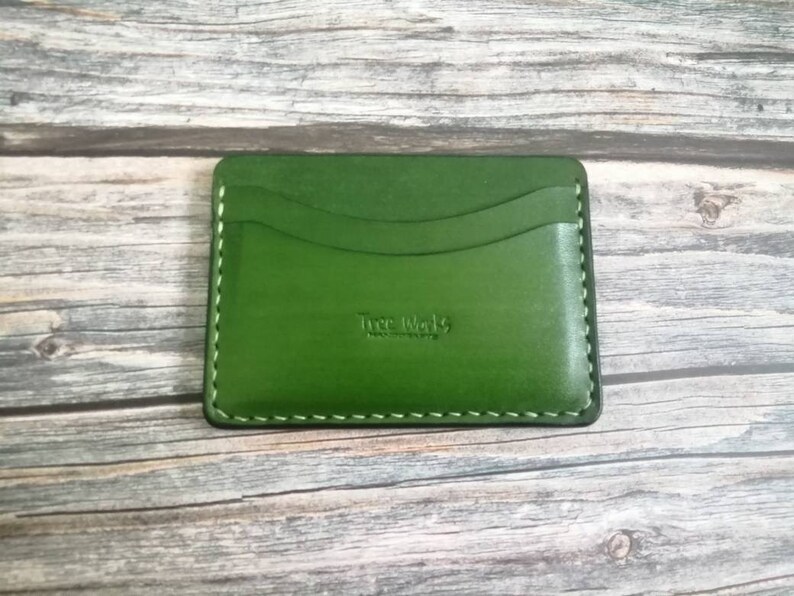 Puede incluir: Un porta tarjetas de cuero verde con un borde cosido y el texto "True Wallets" en relieve en la parte delantera.