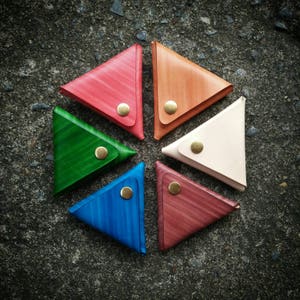 Puede incluir: Seis monederos triangulares de cuero en diferentes colores: rojo, naranja, verde, azul, rosa y blanco. Cada monedero tiene un cierre de presión de latón.