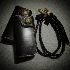 K&ouml;nnte beinhalten: Eine schwarze Ledergeldb&ouml;rse mit einem geflochtenen Lederriemen und zwei Metall-Indianerkopf-Designs. Die Geldb&ouml;rse hat ein Baumdesign auf der Vorderseite.