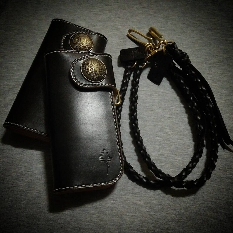 K&ouml;nnte beinhalten: Eine schwarze Ledergeldb&ouml;rse mit einem geflochtenen Lederriemen und zwei Metall-Indianerkopf-Designs. Die Geldb&ouml;rse hat ein Baumdesign auf der Vorderseite.
