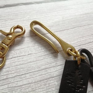 May include: A brass swivel lobster clasp and a brass hook fastener attached to dark brown leather straps. The metal hardware features a polished gold-tone finish, and the leather components are secured with matching brass rings and a small leather wrap.