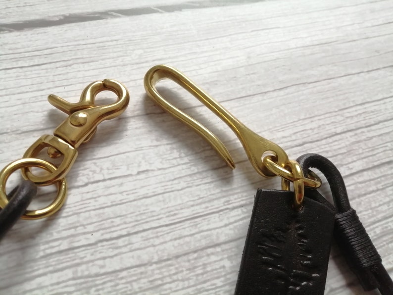 May include: A brass swivel lobster clasp and a brass hook fastener attached to dark brown leather straps. The metal hardware features a polished gold-tone finish, and the leather components are secured with matching brass rings and a small leather wrap.