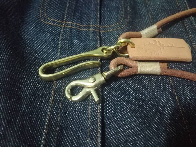 Puede incluir: Primer plano de un llavero de cuero con herrajes de lat&oacute;n. Cuenta con una correa de cuero marr&oacute;n, un gancho de lat&oacute;n y una peque&ntilde;a etiqueta de cuero con un dise&ntilde;o en relieve. El fondo es de tela vaquera azul oscuro.