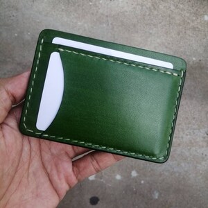 Puede incluir: Una cartera de cuero verde con una ranura para tarjeta blanca y costuras blancas.