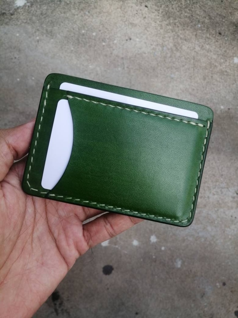 Puede incluir: Una cartera de cuero verde con una ranura para tarjeta blanca y costuras blancas.