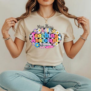 May include: A beige t-shirt with the words "Miss Maria Teaching Assistant" in a colourful, retro design. The design includes flowers, a checkerboard pattern, and school-themed elements. The person is wearing blue jeans.