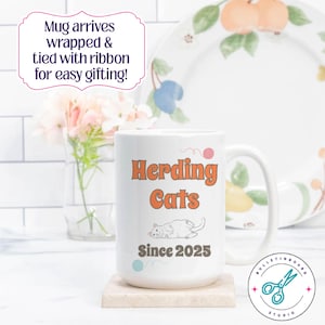 May include: White ceramic mug with the text "Herding Cats Since 2025" in orange and black. The mug features a cartoon cat illustration and is on a small, square, beige coaster. The mug is part of the Bulletinboard Studio collection.