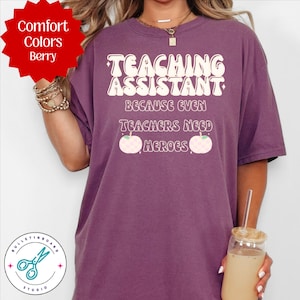 Puede incluir: Camiseta Comfort Colors color bayas con el texto "TEACHING ASSISTANT BECAUSE EVEN TEACHERS NEED HEROES" en blanco. La camiseta presenta una fuente retro y dos gráficos de manzanas. Una etiqueta roja en forma de estrella dice "Comfort Colors Berry".
