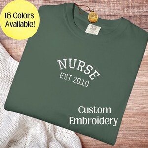 Peut inclure: Un t-shirt vert foncé avec le texte brodé blanc "NURSE EST 2010". Le t-shirt porte également les mots "Custom Embroidery" en blanc. Un cercle jaune dans le coin supérieur gauche indique "16 couleurs disponibles !"