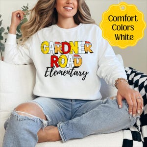 May include: White long-sleeved sweatshirt featuring the text "GARDNER ROAD Elementary" in colourful block letters. A yellow tag in the upper right corner reads "Comfort Colors White". The sweatshirt is worn with ripped blue jeans.