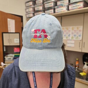 May include: A light blue denim baseball cap with the text "TA Teaching Assistant! Miss De" embroidered in red, yellow, and white.