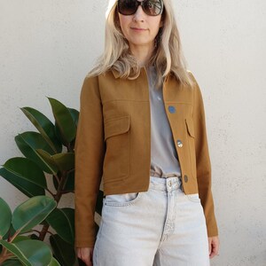 Peut inclure: Une veste courte jaune moutarde avec un col, des manches longues et deux poches poitrine, portée sur un t-shirt gris clair uni. La tenue est complétée par un jean en denim délavé clair à taille haute, coupe large et braguette à boutons.