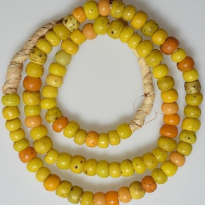 Könnte beinhalten: Eine Halskette aus gelben und orangenen Perlen, auf einem Naturfaserschnur aufgefädelt. Die zylindrischen Perlen weisen unterschiedliche Farbtöne und gesprenkelte Muster auf. Die Halskette ist spiralförmig angeordnet und betont ihr Design.