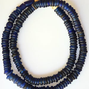 May include: A strand of small, circular, deep blue glass beads strung together. The beads have a slightly irregular, flattened shape and are arranged in a continuous loop, secured at the top with a light-coloured, fibrous cord tied in a knot.