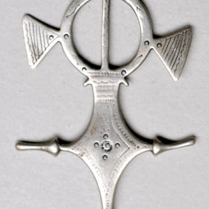 May include: A silver-colored pendant with an intricate design. The pendant features a central vertical bar bisecting a circle, with triangular and wing-shaped elements. The bottom has a diamond shape with a spiral.