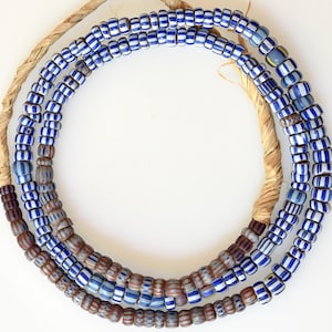Puede incluir: Un collar circular de pequeñas cuentas cilíndricas ensartadas en un cordón de fibra natural de color tostado. Las cuentas presentan rayas verticales alternas de blanco opaco y azul profundo, intercaladas con secciones de cuentas marrones y grises.