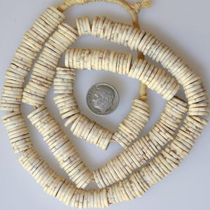 May include: A long strand of off-white, disc-shaped beads with mottled brown speckles, arranged in a spiral pattern on a string. A silver-colored United States dime is placed in the center for scale, showing the relative size of the small, flat beads.