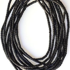Puede incluir: Una hebra de cuentas de vidrio negro, cada cuenta es cilíndrica y ligeramente redondeada. Las cuentas están enhebradas en un cordón de fibra natural.