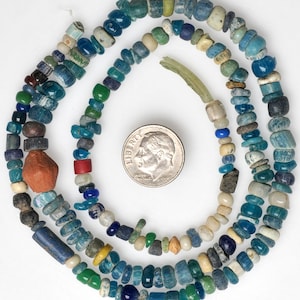 Puede incluir: Una colección de cuentas de estilo antiguo en varios tonos de azul, verde y blanco, dispuestas en un patrón circular alrededor de una moneda plateada para mostrar la escala. Las cuentas varían en forma, tamaño y textura, incluyendo una cuenta naranja grande.