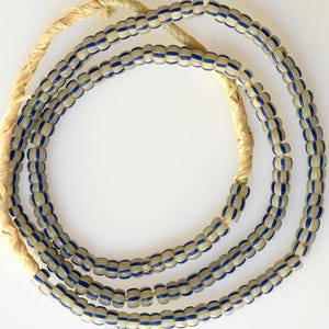 Puede incluir: Un collar de cuentas color crema con rayas azules, ensartadas en un cordón de fibra natural. Las cuentas cilíndricas están espaciadas uniformemente, creando un patrón repetitivo. El cordón es de color beige claro.