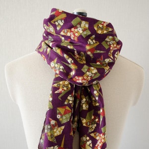 May include: A purple scarf with a pattern of origami cranes in shades of green, gold, and red. The scarf has a fringed edge.
