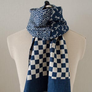 May include: A blue and white patterned scarf displayed on a mannequin. The scarf features a geometric design, dragonfly motifs, and a checkered pattern. The scarf has a fringed edge.