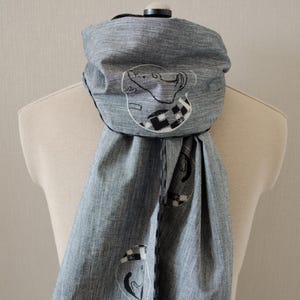 May include: A gray scarf with black trim and a white and black embroidered design of a cat. The cat is wearing a hat with a checkered pattern.