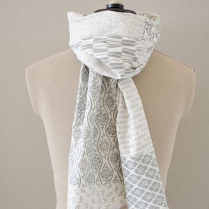 May include: A white and gray patterned scarf draped over a mannequin. The scarf features various geometric and floral designs in gray against a white background. The scarf's edges have a fringed detail.