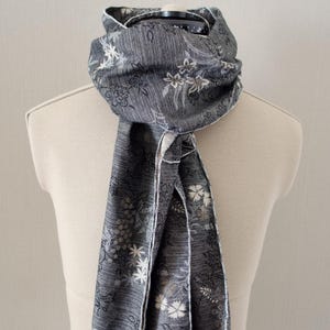 May include: A gray scarf with a white floral pattern. The scarf has a fringed edge and is made of a soft, textured fabric.