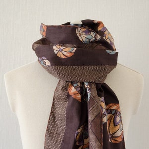 May include: A dark brown scarf with a repeating pattern of colorful, stylized floral designs. The scarf has a textured border and a fringed edge. The scarf is draped over a mannequin.