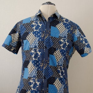 Könnte beinhalten: Ein blaues und weißes kurzärmeliges Hemd mit Knöpfen und Patchwork-Muster. Das Hemd hat verschiedene geometrische Muster in Blautönen und Weiß.