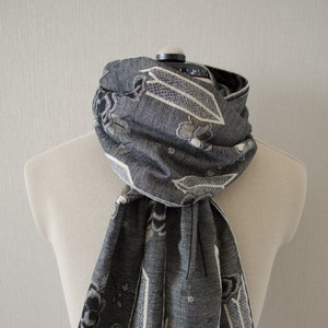 May include: A gray scarf with a floral pattern and fringed edges is draped over a mannequin. The scarf features a repeating design of flowers and geometric shapes in shades of gray, black, and white.