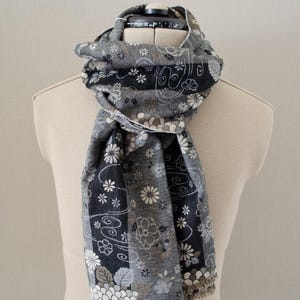 May include: A gray scarf with a floral pattern, displayed on a mannequin. The scarf features a mix of white, black, and gray flowers and foliage. The edges have a fringed trim. The scarf is wrapped around the mannequin's neck.