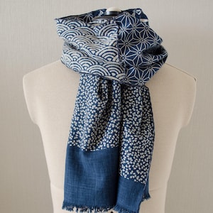 May include: A blue and white patterned scarf draped over a mannequin. The scarf features various designs, including wave patterns, geometric shapes, and small floral prints. The bottom edge has a fringed trim.