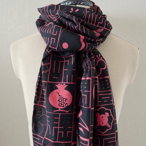 May include: Black scarf with a repeating pattern of pink geometric shapes and symbols. The scarf is draped over a mannequin. The design includes a pomegranate, yin-yang symbol, and other abstract elements. The scarf has a fringed edge.