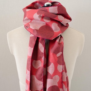 May include: A red scarf featuring a pattern of large, light pink circles. The scarf is draped over a mannequin, highlighting its design and texture. The scarf's edges are finished with a darker pink trim.