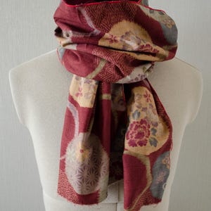 May include: A burgundy scarf with a floral and geometric pattern, draped over a mannequin. The scarf features a deep red interior and a decorative edge. The design includes shades of beige, gray, and burgundy, creating a visually rich textile.