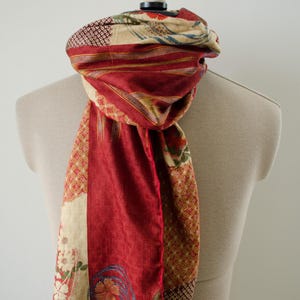 Op de afbeelding: Een rechthoekige sjaal met een patchworkontwerp in dieprode, crèmekleurige en bruine tinten. De stof toont een mix van bloemmotieven, geometrische patronen en dunne strepen, met een zachte, getextureerde afwerking en gerafelde randen aan de onderkant.