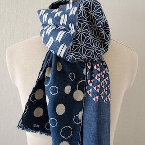 May include: A blue and white patterned scarf draped over a mannequin. The scarf features various designs, including white brushstrokes, geometric shapes, and polka dots. The edges are frayed, adding a textured detail.