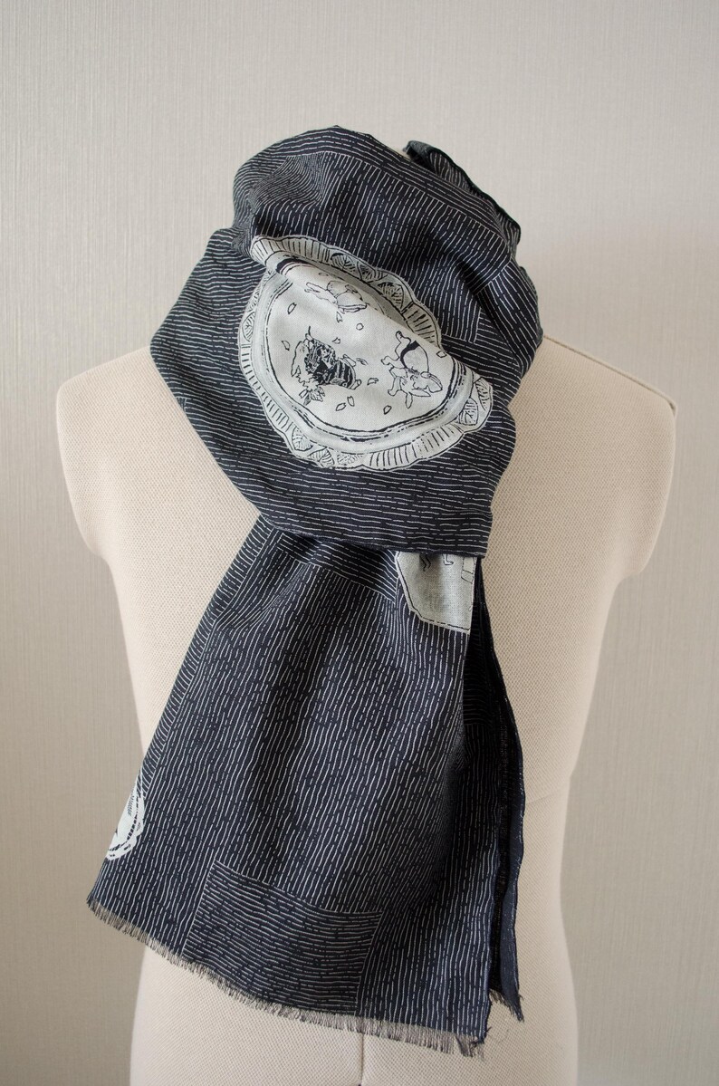 May include: Dark blue scarf with white vertical stripes and a decorative circular patch featuring a whimsical illustration. The scarf is draped over a mannequin, showcasing its design and texture. The fabric appears to be soft and lightweight.