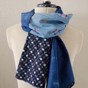 May include: A blue scarf with a unique design is draped over a mannequin. The scarf features a combination of solid blue fabric, a light blue section with red stripes, and a dark blue patterned section with white and red accents. The scarf is made of soft fabric.