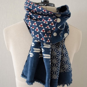 May include: A blue scarf with various geometric patterns in white and red, draped over a mannequin. The scarf features triangles, circles, and starburst designs. The edges have a fringed trim.