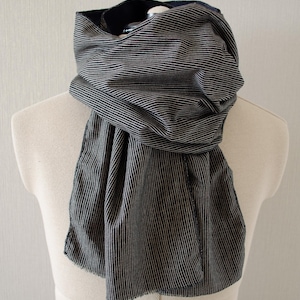 May include: A striped scarf in shades of gray and white is draped over a mannequin. The scarf has a classic design with vertical stripes. The fabric appears soft and lightweight, suitable for cooler weather. The scarf is a versatile accessory.