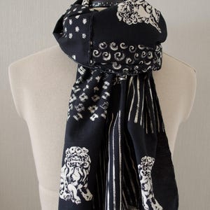 May include: Black and white patterned scarf with a variety of designs, including lion figures, stripes, and swirls. The scarf is draped over a mannequin and has a fringed edge.