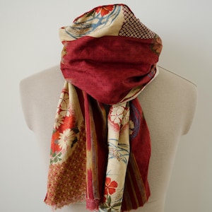 May include: A rectangular scarf featuring a deep red and cream color palette with intricate floral patterns, geometric shapes, and striped sections. The fabric has a textured appearance with frayed edges, draped around a neutral-toned mannequin.