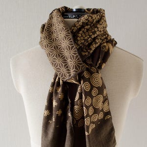 May include: A brown scarf with various geometric and floral patterns in a lighter tan color. The scarf is draped over a mannequin, showcasing the different designs, including spirals, starbursts, and small flower clusters. The scarf has a fringed edge.